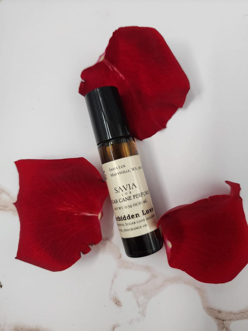 May include: A roll-on perfume bottle with a black cap and a label that reads "SAVIA LUX Sugar Cane Perfume Forbidden Love." The bottle is surrounded by three bright red rose petals on a white and gray marble surface.