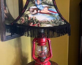 May include: A red vintage-style oil lamp with a glass globe and a black fringed lampshade featuring a pin-up style illustration of a woman in a forest setting.