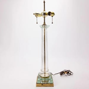 May include: A clear glass column lamp with brass accents. The lamp features a rectangular base and a clear glass column. The top has two light sockets with pull chains. The lamp is a classic design.