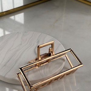 Puede incluir: Pinza para el pelo de metal dorado con un marco rectangular y un cierre de resorte. El marco tiene un patrón decorativo.