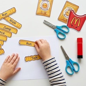 May include: A set of paper cutouts featuring cartoon condiment packets, a fry container, and crinkle-cut french fry shapes labelled with action verbs like Run, Walk, Jump, Dance, Sleep, Sit, Stand, and Talk, alongside blue scissors and a red glue stick.