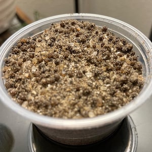 May include: A clear plastic container filled with a mixture of small, granular, and crystalline substances. The contents appear to be a light brown color with translucent, off-white crystals interspersed throughout. The container is sitting on a dark surface.