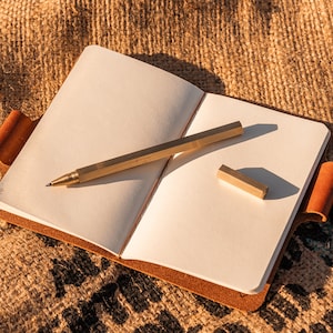 K&ouml;nnte beinhalten: Ein leeres, wei&szlig;es Notizbuch mit braunem Lederumschlag und einem Messing-Stift und -Lineal darauf. Das Notizbuch liegt auf einer Jutestoff-Oberfl&auml;che.