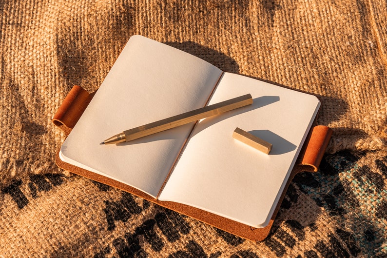 K&ouml;nnte beinhalten: Ein leeres, wei&szlig;es Notizbuch mit braunem Lederumschlag und einem Messing-Stift und -Lineal darauf. Das Notizbuch liegt auf einer Jutestoff-Oberfl&auml;che.