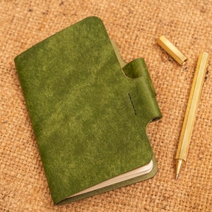 K&ouml;nnte beinhalten: Ein gr&uuml;nes, ledergebundenes Notizbuch mit einer Klappe, das auf einer Jutestoffoberfl&auml;che liegt, neben einem goldenen Stift.