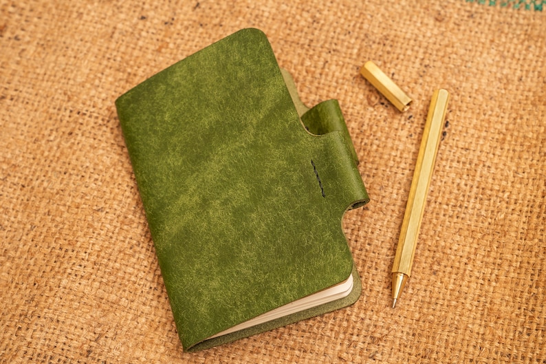 K&ouml;nnte beinhalten: Ein gr&uuml;nes, ledergebundenes Notizbuch mit einer Klappe, das auf einer Jutestoffoberfl&auml;che liegt, neben einem goldenen Stift.