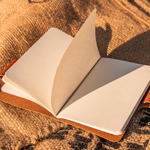 K&ouml;nnte beinhalten: Ein braunes, ledergebundenes Tagebuch mit leeren Seiten liegt offen auf einer Jutestoff-Oberfl&auml;che. Das Tagebuch hat einen braunen Lederriemen auf der rechten Seite.