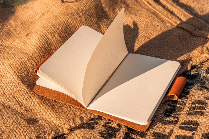 K&ouml;nnte beinhalten: Ein braunes, ledergebundenes Tagebuch mit leeren Seiten liegt offen auf einer Jutestoff-Oberfl&auml;che. Das Tagebuch hat einen braunen Lederriemen auf der rechten Seite.