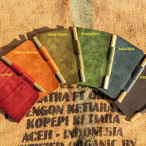K&ouml;nnte beinhalten: Sechs ledergebundene Notizb&uuml;cher in verschiedenen Farben, darunter Bordeauxrot, Cognacbraun, Olivgr&uuml;n, Evergreen, Aquablau und Mitternachtsblau. Jedes Notizbuch hat eine Stiftschlaufe aus Messing und einen Messingverschluss.