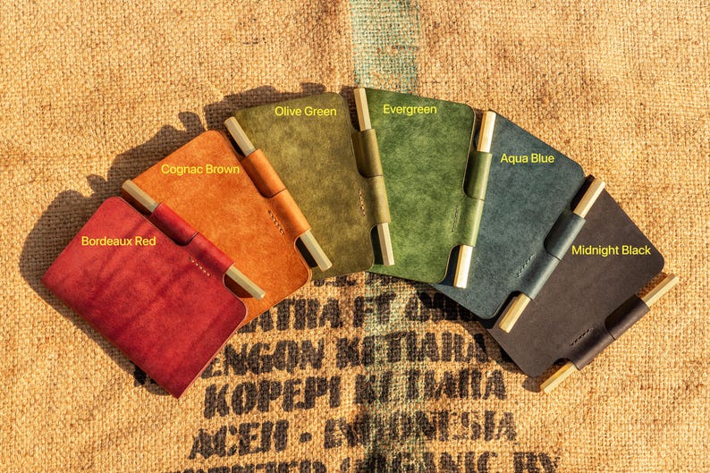K&ouml;nnte beinhalten: Sechs ledergebundene Notizb&uuml;cher in verschiedenen Farben, darunter Bordeauxrot, Cognacbraun, Olivgr&uuml;n, Evergreen, Aquablau und Mitternachtsblau. Jedes Notizbuch hat eine Stiftschlaufe aus Messing und einen Messingverschluss.