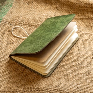 以下が含まれることがあります： クリーム色のページと白いゴム留めが付いた、開いたオリーブグリーンの革製ジャーナル。ジャーナルは、質感のあるタン色のジュートの表面に置かれています。表紙はわずかに開いており、ページが見えています。