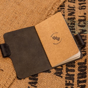 K&ouml;nnte beinhalten: Ein schwarzes Ledernotizbuch mit einem braunen Papierumschlag. Der Umschlag tr&auml;gt ein Logo mit der Aufschrift "Marple Lake" und "Handcrafted Goods".