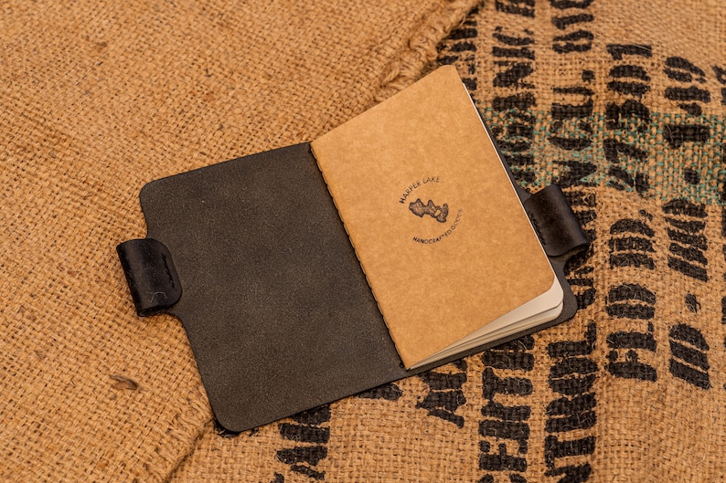 K&ouml;nnte beinhalten: Ein schwarzes Ledernotizbuch mit einem braunen Papierumschlag. Der Umschlag tr&auml;gt ein Logo mit der Aufschrift "Marple Lake" und "Handcrafted Goods".