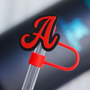 Può includere: Un copripaglie a forma di lettera A rossa e nera, realizzato in materiale flessibile. L'accessorio presenta una A stilizzata con centro rosso e bordo nero, fissato a un anello rosso che lo tiene fermo su una cannuccia di plastica trasparente.