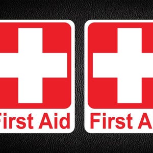 May include: Two red and white first aid stickers. Each sticker features a white cross on a red background, with the words "First Aid" in white text below. The stickers are on a black textured surface.