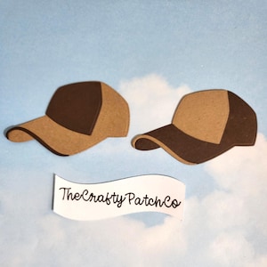 May include: Two brown and tan baseball cap cutouts against a light blue background. The caps are side-by-side, with the text "The Crafty Patch Co" below them on a white banner.