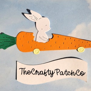 May include: A white bunny rabbit sitting in a carrot shaped car with green leaves and yellow wheels. The car has a bite taken out of it. The text "The Crafty Patch Co." is on a white banner below the car.
