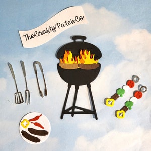 May include: A selection of barbecue-themed paper cut-outs. Features a black grill with flames, a plate of food, skewers, and grilling tools. The text "The Crafty Patch Co" is displayed on a banner.