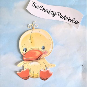 May include: A yellow duck with orange feet and beak, sitting on a light blue background. The duck has a stitched design and a small pink blush on its cheek. The text "The Crafty Patch Co." is visible in the background.