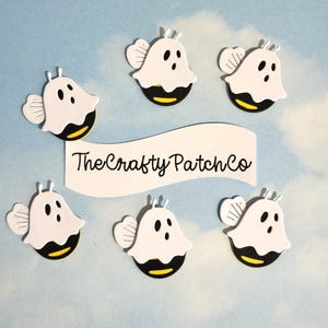 May include: Six white bee-shaped embellishments with black eyes and a yellow and black striped bottom. The embellishments have white wings and antennae. The text "The Crafty Patch Co" is on a white banner.