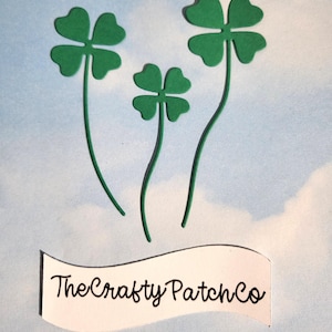 Könnte beinhalten: Drei grüne vierblättrige Kleeblätter mit gebogenen Stielen vor einem hellblauen Hintergrund mit weißen wolkenartigen Formen. Ein weißes Banner am unteren Rand zeigt den Text "The Crafty Patch Co" in schwarzer Schreibschrift.
