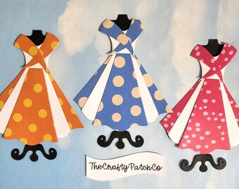 Könnte beinhalten: Drei Papierkleid-Ausschnitte auf schwarzen Ständern mit orangefarbenen, blauen und rosafarbenen Punkten sowie weißen Paneelen. Jedes Kleid hat ein überkreuztes Oberteil. Ein weißes Banner am unteren Rand zeigt den Text TheCraftyPatchCo.
