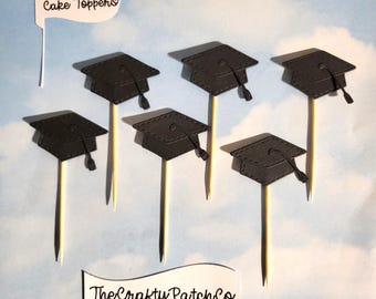 May include: Graduation-themed cake toppers. The toppers feature black mortarboard caps attached to wooden sticks. A white banner with the text "Cake Toppers" is visible, along with a banner that reads "The Crafty Patch Co."