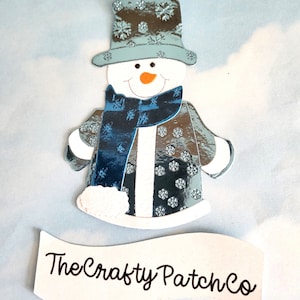 Può includere: Un ritaglio di pupazzo di neve stravagante con cappello, sciarpa e cappotto blu e argento, adornato con motivi a fiocco di neve. Il pupazzo di neve ha un naso a carota e un viso sorridente. Lo striscione sottostante recita "The Crafty Patch Co."