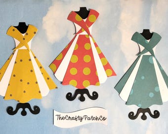 Könnte beinhalten: Drei Papierkleider in Gelb, Rot und Türkis, jedes mit einem einzigartigen Tupfenmuster. Die Kleider werden auf schwarzen Ständern vor einem hellblauen Hintergrund präsentiert. Das Logo The Crafty Patch Co. ist sichtbar.