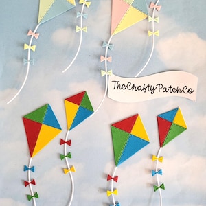 Op de afbeelding: Handgemaakte vliegers in pastel- en primaire kleuren, elk met een uniek ontwerp. De vliegers zijn versierd met kleine strikaccenten en bevestigd aan witte touwtjes. De achtergrond is een lichtblauwe lucht met wolken.