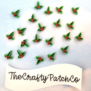 May include: A collection of twenty-one small, three-dimensional holly leaf and berry embellishments, each featuring green leaves and three red berries. Below the holly pieces is a white banner with the black text The Crafty Patch Co.