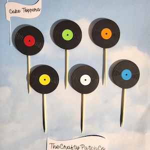 Op de afbeelding: Zes zwarte vinylplaat taarttoppers met gekleurde middenlabels: rood, groen, oranje, geel, wit en blauw. Elke plaat is bevestigd aan een houten stok. De tekst "Cake Toppers" en "The Crafty Patch Co" zijn ook zichtbaar.