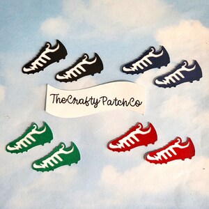 May include: A selection of football boots in black, navy, green, and red with white detailing. The boots are displayed against a light blue, cloudy background. The text "The Crafty Patch Co" is visible on a white banner.