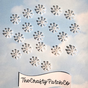 Puede incluir: Una colección de pequeños adornos blancos con forma de copo de nieve hechos de diminutas perlas artificiales dispuestas en un patrón radial. Debajo de los copos hay una pancarta de papel blanco con el texto negro TheCraftyPatchCo.