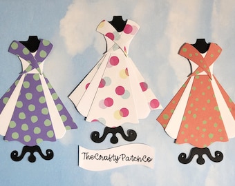 Könnte beinhalten: Drei Papierkleider in Lila, Weiß und Orange, jedes mit Tupfen, stehen auf schwarzen Ständern vor einem blauen Himmel. Die Kleider stehen auf schwarzen Ständern. The Crafty Patch Co. steht auf einem weißen Banner.