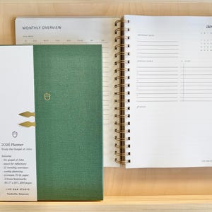 Könnte beinhalten: Ein grüner 2026-Planer mit goldener Spiralbindung und weißem Rücken. Der Planer enthält eine monatliche Übersicht und eine Januar-Kalenderseite. Das Cover zeigt ein Eicheldesign und den Text "2026 Planner".