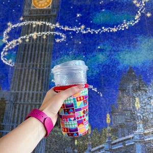 May include: A clear plastic cup with a red-rimmed neoprene sleeve featuring a colorful geometric pattern of squares, triangles, and circles in shades of blue, purple, yellow, and red, topped with a clear plastic lid.