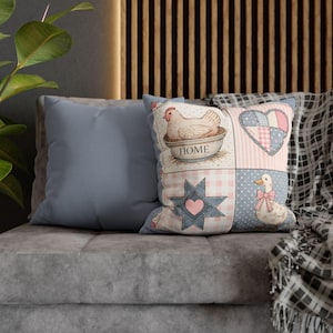 May include: Two decorative throw pillows on a gray sofa. One pillow is solid light blue. The other pillow has a patchwork design with a hen, heart, star, and duck. The word "HOME" is printed on the pillow.