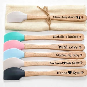 Może przedstawiać: Zestaw sześciu silikonowych szpatułek z jasnymi drewnianymi uchwytami. Każdy uchwyt jest wygrawerowany z niestandardowym tekstem, w tym Olivia's baby shower, Michelle's kitchen, With Love, Welcome my Baby, Love is sweet Ruby & Scott oraz Kenna & Ryan.