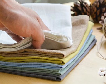 May include: A stack of folded fabric napkins in various colors, including white, beige, yellow, blue, and gray. The napkins are arranged in a neat pile on a wooden surface.