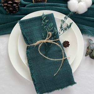 Könnte beinhalten: Eine gefaltete, teal-farbene Leinen-Serviette mit ausgefransten Kanten, mit Bindfaden gebunden und mit Zweigen aus getrocknetem Grün geschmückt, liegt auf einem weißen Teller. Ein Tannenzapfen und ein Baumwollballen sind im Hintergrund zu sehen.