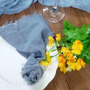Könnte beinhalten: Eine Tischdekoration mit einer blauen Leinen-Serviette mit ausgefransten Kanten, einem weißen Teller, einem Weinglas und gelben Blumen. Ein blauer Stoffläufer ist über eine Holzoberfläche drapiert und schafft eine rustikale und elegante Ästhetik.