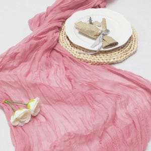 May include: A dusty rose-colored, crinkled fabric table runner with a white plate, woven placemat, and rolled paper napkin tied with a silver ribbon. Two white faux flowers are placed on the runner.