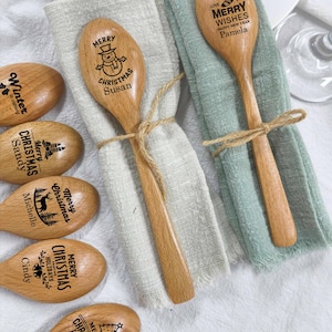 Könnte beinhalten: Eine Sammlung hellbrauner Holzlöffel mit lasergravierten Weihnachtsmotiven und Namen wie Susan, Pamela, Sandy, Michelle, Cindy und Isidora. Zwei Löffel sind mit Bindfaden an beigen und salbeigrünen Stoffservietten befestigt.