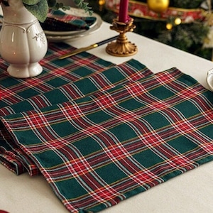 Puede incluir: Un camino de mesa de cuadros verdes y rojos con fondo blanco. El camino de mesa está doblado por la mitad y está sobre una mesa blanca. Hay una vela roja en el centro del camino de mesa.