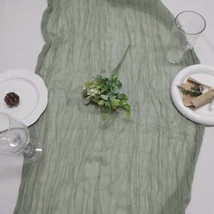 Könnte beinhalten: Eine Tischdekoration mit einem strukturierten, salbeigrünen Tischläufer, weißen Tellern, klaren Gläsern und einem Zweig Grün. Ein Teller hat einen Tannenzapfen, der andere eine Schleife aus Jute. Die Szene ist auf einer weißen Oberfläche.
