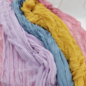 Könnte beinhalten: Eine Sammlung farbenfroher, leichter Schals in Rosa-, Lavendel-, Blau- und Gelbtönen. Die Schals bestehen aus einem weichen, fließenden Stoff mit gekreppter Textur. Die Schals sind in einem geschichteten, diagonalen Muster angeordnet.