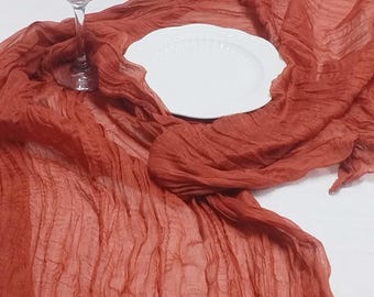Könnte beinhalten: Ein klares Weinglas und ein weißer Teller liegen auf einem rostfarbenen, zerknitterten Stoff-Tischläufer. Der Läufer drapiert sich über eine weiße Oberfläche und erzeugt eine strukturierte und elegante Präsentation. Ideal für Hochzeiten oder besondere Anlässe.