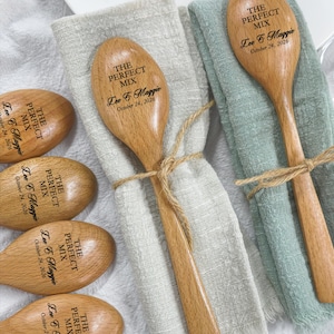 May include: A collection of light brown wooden spoons, each engraved with the text THE PERFECT MIX, Lee & Maggie, and October 24, 2026. Two spoons are tied with twine to beige and sage green linen napkins, while others are arranged nearby.
