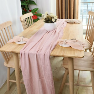 Puede incluir: Un camino de mesa rosa claro con bordes deshilachados, colocado sobre una mesa de madera con dos sillas. Un jarrón blanco con flores blancas se encuentra sobre el camino de mesa.